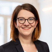 Smiling, Caucasian woman with reddish brown hair wearing round spectacles and a black blazer jacket