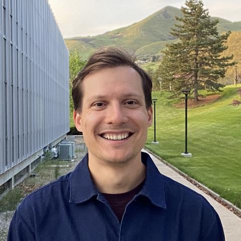 A professional headshot of Max wearing a dark blue shirt. He is smiling and standing outdoors with rolling hills in the background, suggesting a campus setting.