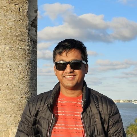 Naimul is wearing sunglasses, a black puffer jacket, and a red patterned shirt is smiling while standing outdoors. 