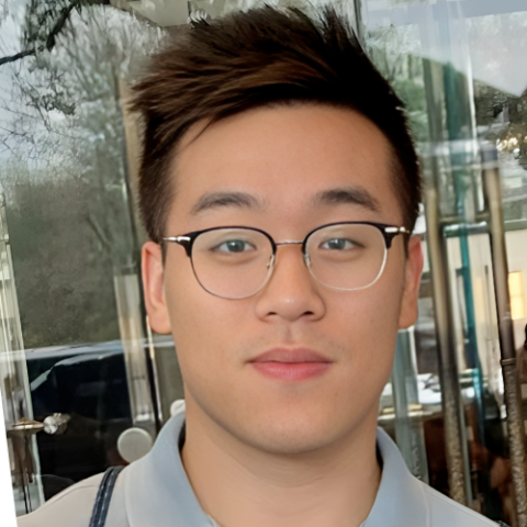 Yu us with short, styled hair and glasses is looking at the camera with a neutral expression. He is wearing a light gray shirt and is standing in front of a glass door with reflections of trees and buildings visible in the background. 