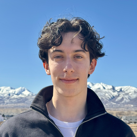 Sam is standing outdoors and is wearing a black jacket over a white shirt. The background features a clear blue sky and snow-capped mountains