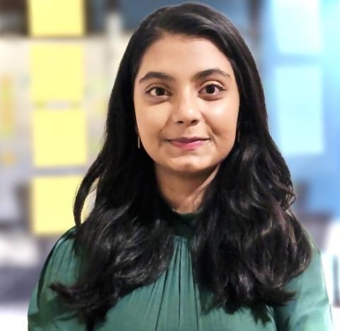 A professional headshot of Srishti, wearing a green top, standing against a blurred background with light and dark blue hues, likely some office workspace.